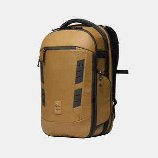 A tan and black backpack shown from a three-quarter angle on a clean white background. The backpack features a tan body with black accents, including zippers, trim, and side panels, and has a small rectangular logo patch on the lower front.