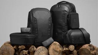 Black NOMATIC camera backpacks, slings, and accessories arranged on logs