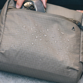 Close-up of a beige waterproof bag with water droplets on the surface, showing a hand holding a smartphone inside the top pocket.