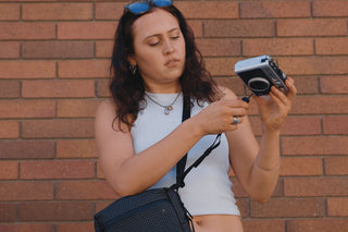 A woman with dark curly hair wearing a white tank top and sunglasses on her head, holding a silver camera and looking down at it. She has a black crossbody bag with a textured pattern and a silver necklace.