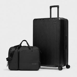 A black hard-shell spinner suitcase with an extended telescoping handle stands next to a matching black duffel bag with top handles and a shoulder strap. The brand name 'NOMATIC' is subtly printed in white on the lower front of the duffel bag.