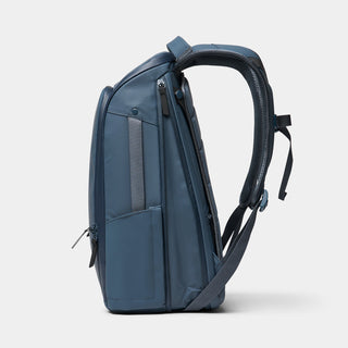 A side profile view of a slate blue backpack with dark gray accents, zippers, and a padded shoulder strap, set against a light gray studio background.