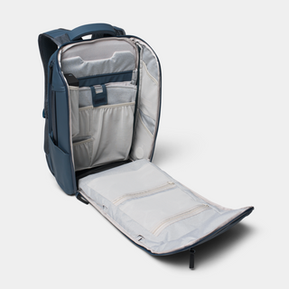 An open navy blue backpack shown from the side, revealing a light gray interior with multiple zip compartments, organizer pockets, and padded sections.