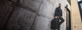 A man in a black long-sleeve shirt and dark pants sits on a concrete wall, holding a black backpack or duffel bag. The photo is taken from a low angle against a textured gray wall, with part of a beige building visible on the right.