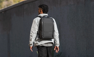 NOMATIC Travel Pack Bundles collection banner, a man walking away from camera wearing a black backpack against a dark textured wall