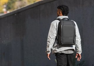 NOMATIC Travel Bag 30L & 40L collection banner, a man walking away from camera wearing a black backpack against a dark textured wall