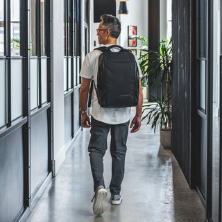 Man wearing a black NOMATIC backpack while walking through a modern office hallway.