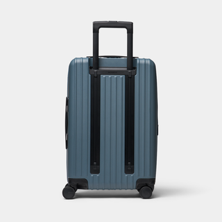 A blue-gray hardshell spinner suitcase with a telescoping handle extended, shown from the back against a light gray studio background.