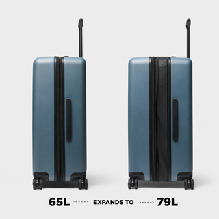 Two hard-shell spinner suitcases shown side by side on a light gray background, with text below reading 65L EXPANDS TO 79L. The left suitcase is shown at 65L capacity and the right at 79L, both in a muted blue color with black zippers, handles, and wheels.