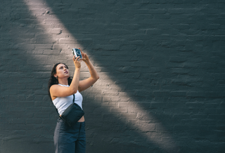 A woman in a white tank top and dark pants takes a photo against a dark gray brick wall with dramatic diagonal sunlight and shadows.