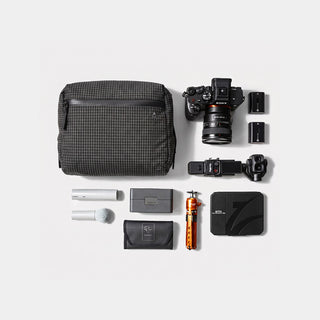 A flat-lay product shot of a camera gear kit on a light gray background, including a black and gray checkered camera bag, a mirrorless camera with lens, battery packs, a camera grip, a small orange accessory, a black pouch, and other matching accessories.