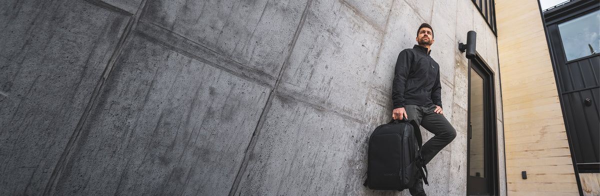 A man in a black long-sleeve shirt and dark pants sits against a large concrete building wall, holding a black briefcase.